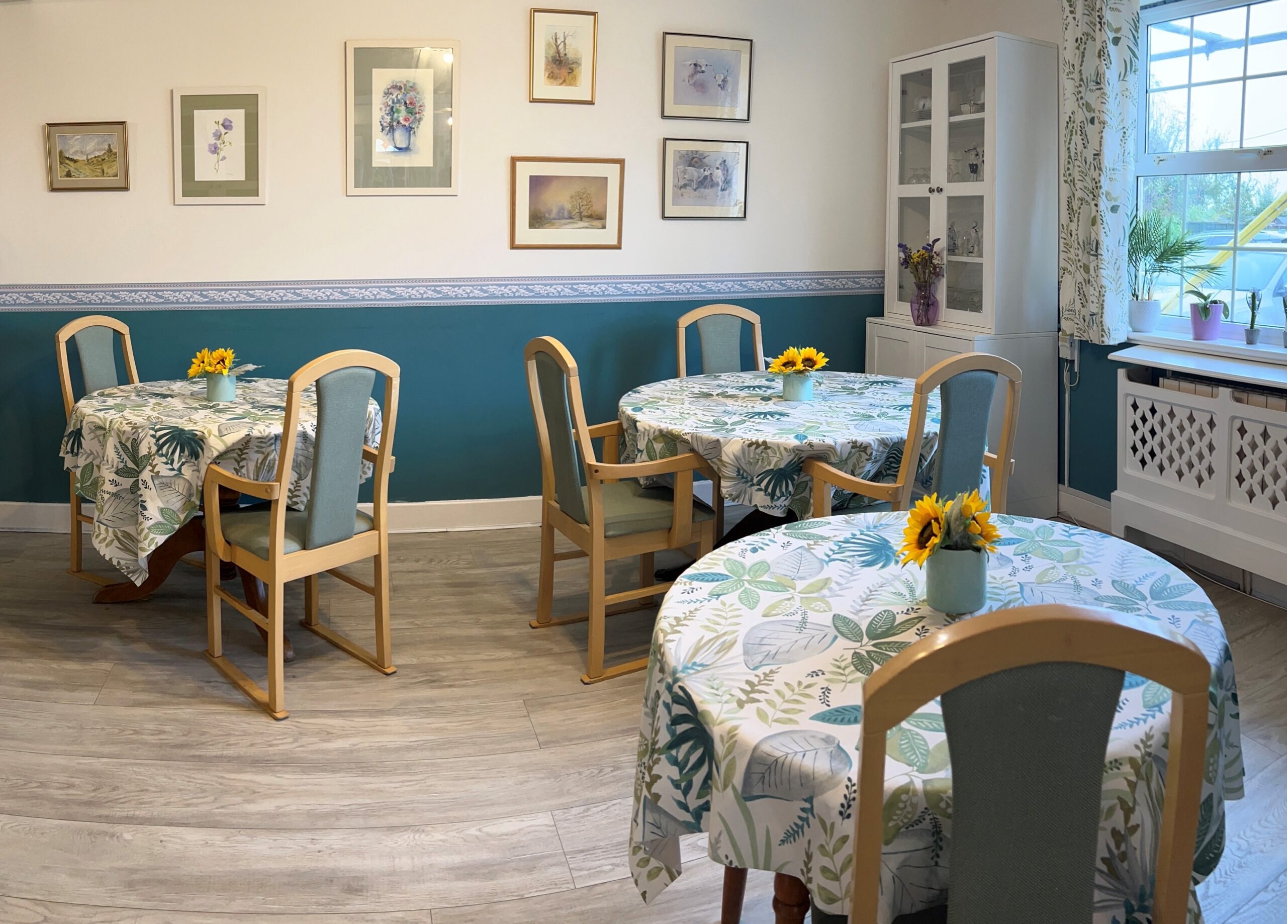 The dining room at Woodfalls Care Home with laid tables, floral tablecloths and framed pictures for the contact Woodfalls Care Home page